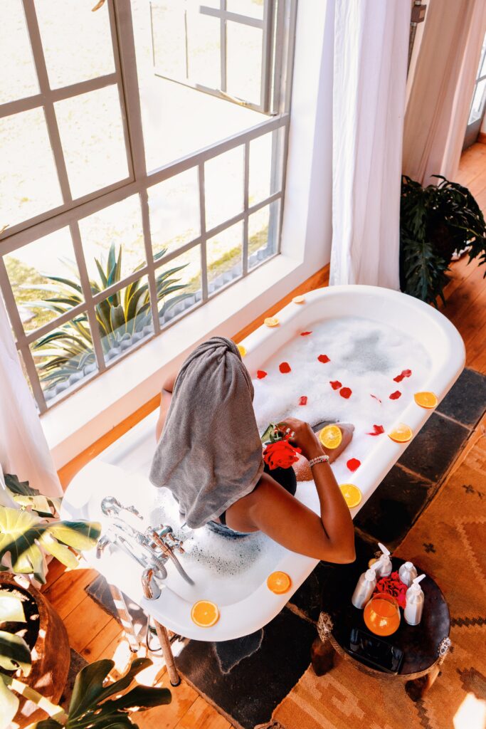 A woman enjoys a relaxing bath with citrus slices and rose petals in a sunlit room.